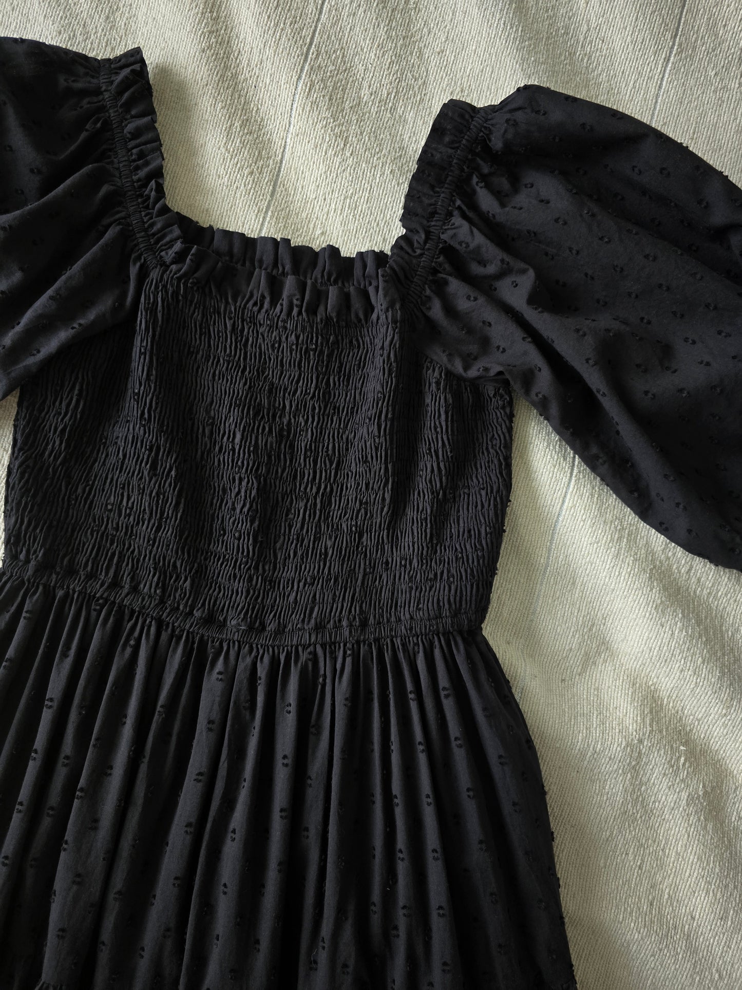 Detailaufnahme des schwarzen Midi-Kleids aus Baumwolle mit Puffärmeln und elastischem Smok-Oberteil.