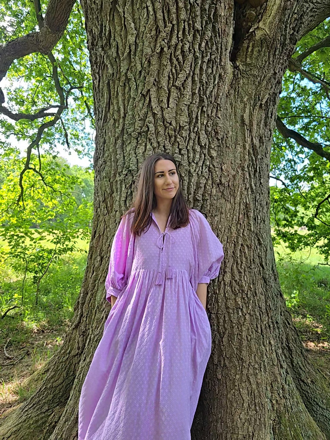 Frau im lila Lucia Midikleid steht entspannt vor einem großen Baum in der Natur.