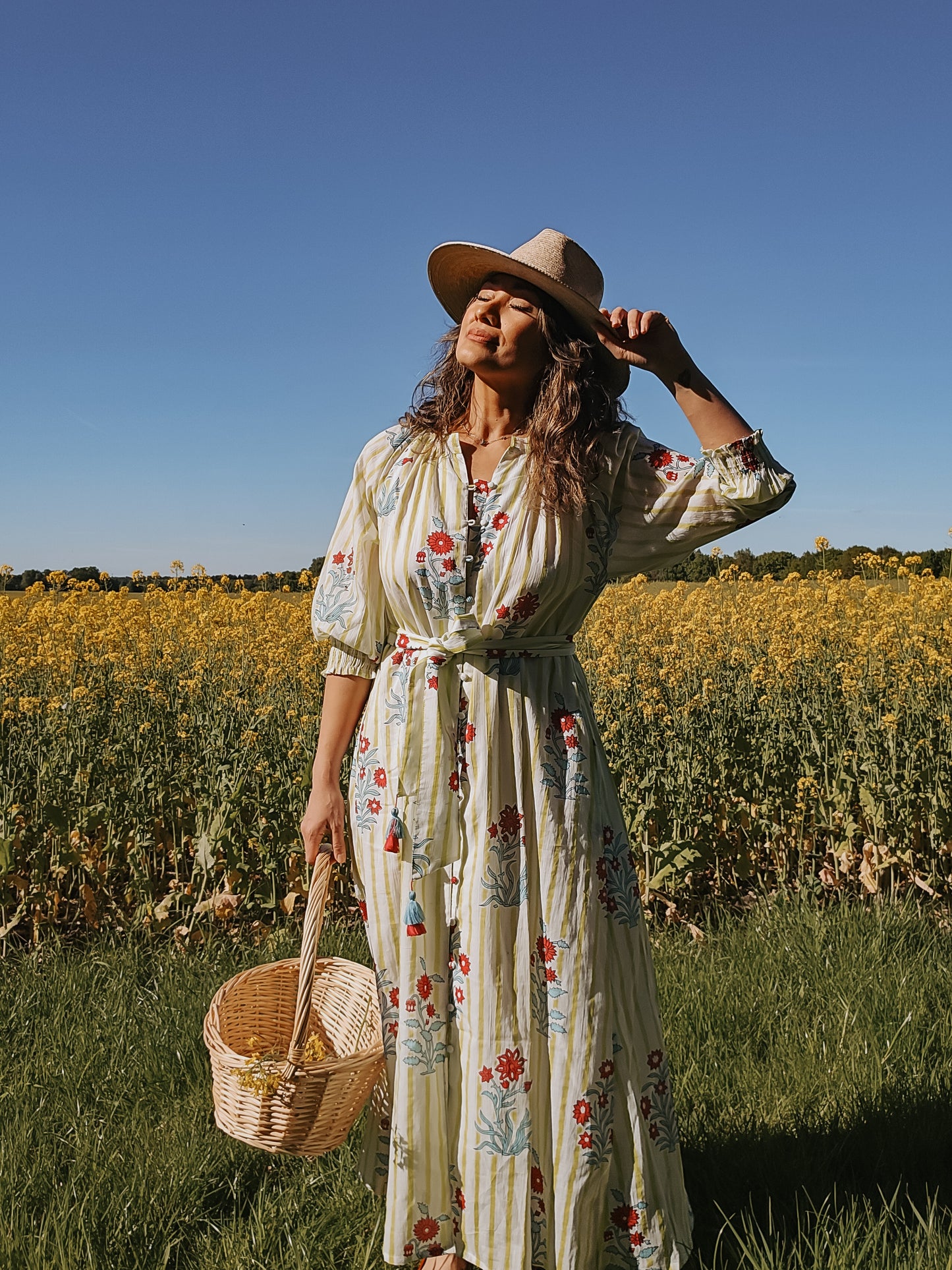 Rumi – Handbedrucktes Baumwollkleid in einem Blumenfeld mit einem Strohhut und einem Korb, handgefertigt im Slow Fashion Stil.
