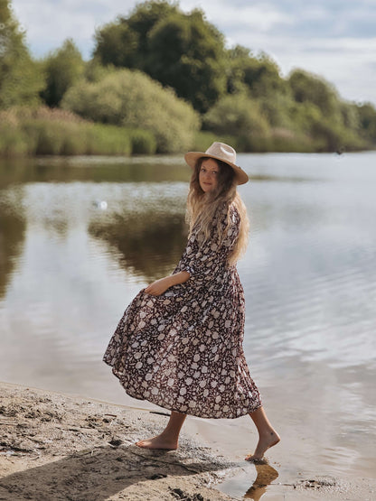 Camilla Kleid mit Handblockdruck – Burgundy, getragen von einer Frau am Wasser mit einem Hut.
