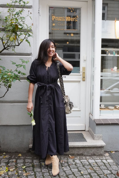 Rumi Kleid - Schwarz: Modell trägt das handgemachte Kleid mit Taschen und Knopfleiste vor einem Galerieeingang.